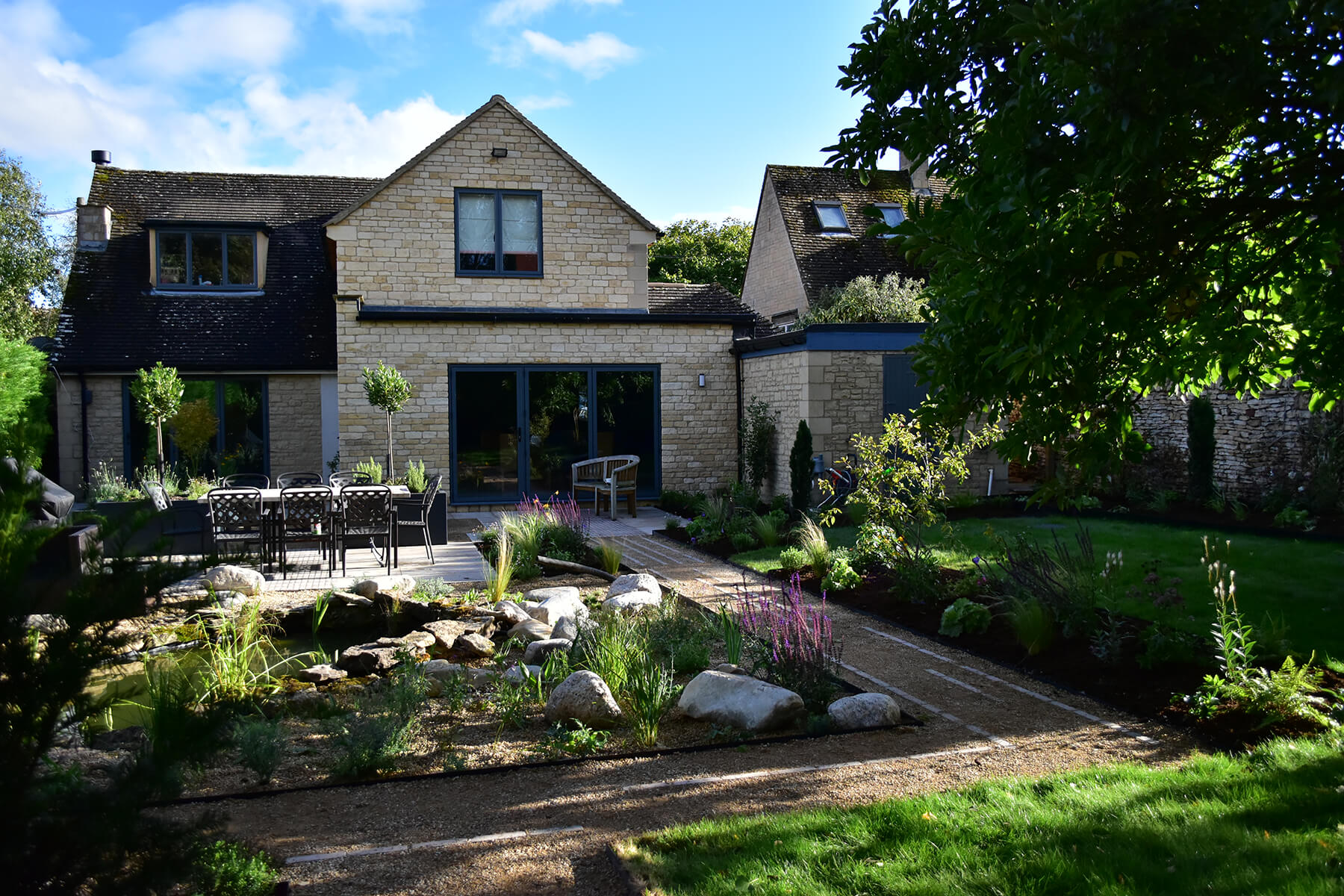 The Pond Garden, a back garden in Donnington, Cotswolds, with a limestone seating terrace, large raised aluminium planters and a fish pond surrounded by seasonal planting. Designed by April House – Award Winning Cotswolds Garden Design